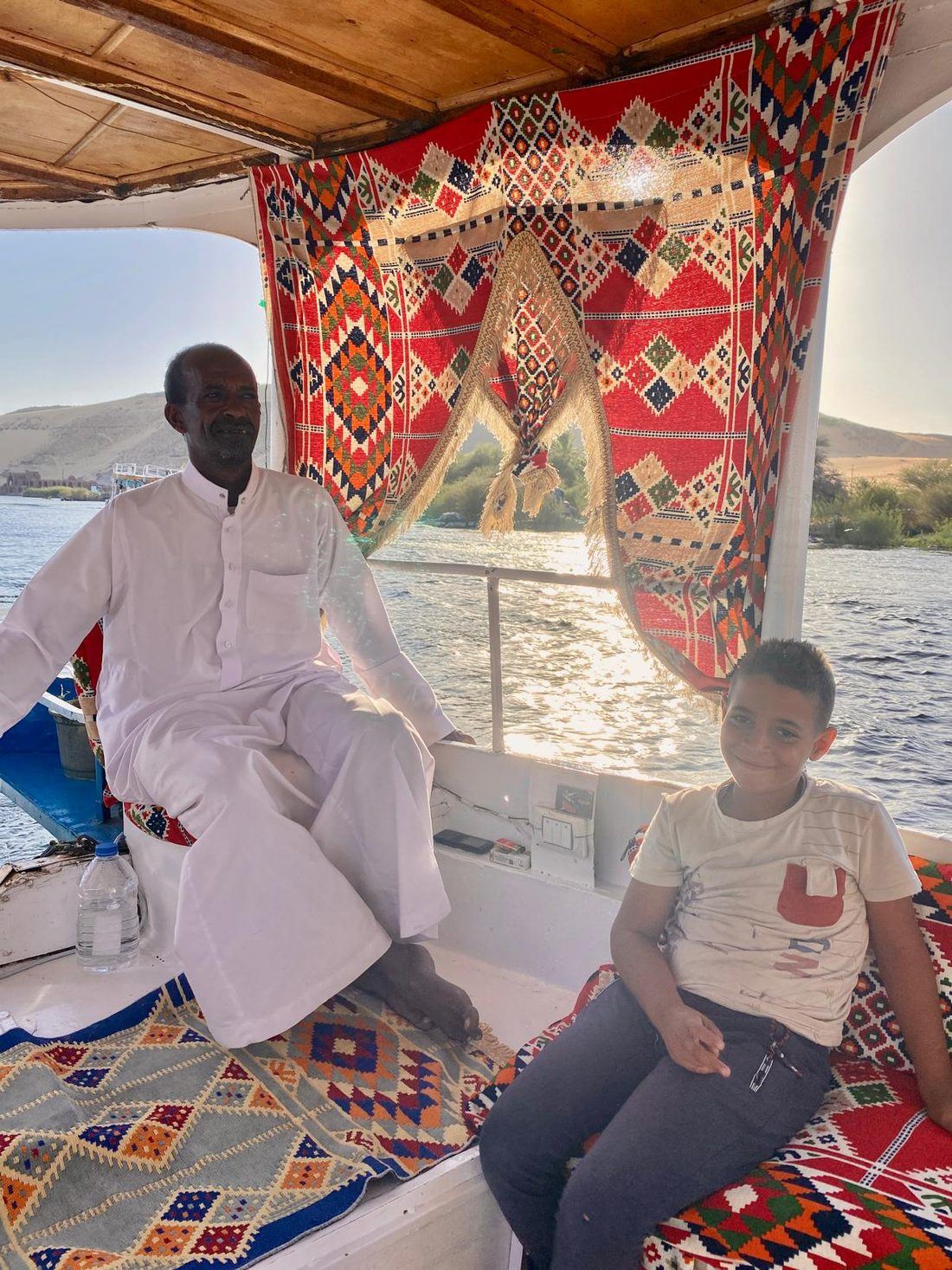A felucca captain in white seated with a boy on patterned cushions under the boat's canopy