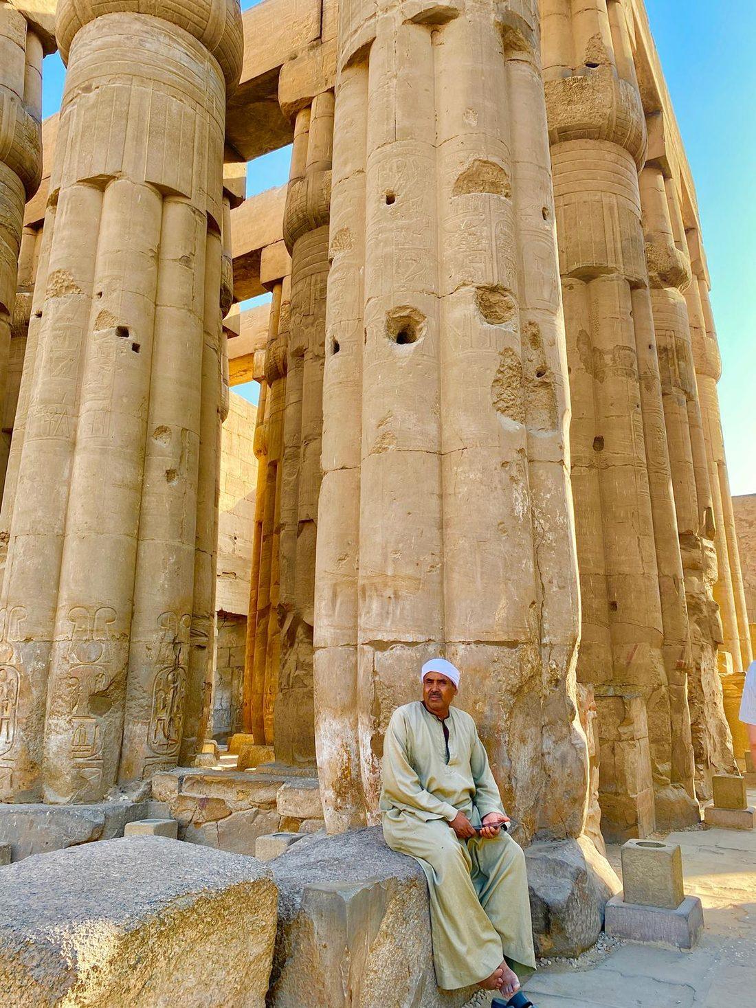 A man seated at the base of the towering carved columns of Luxor Temple