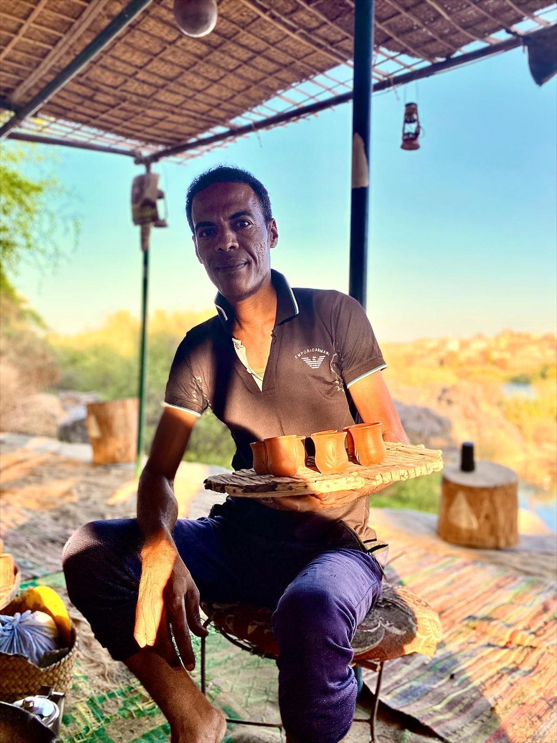 A Nubian host carrying a tray of tea glasses under a reed canopy above the Nile